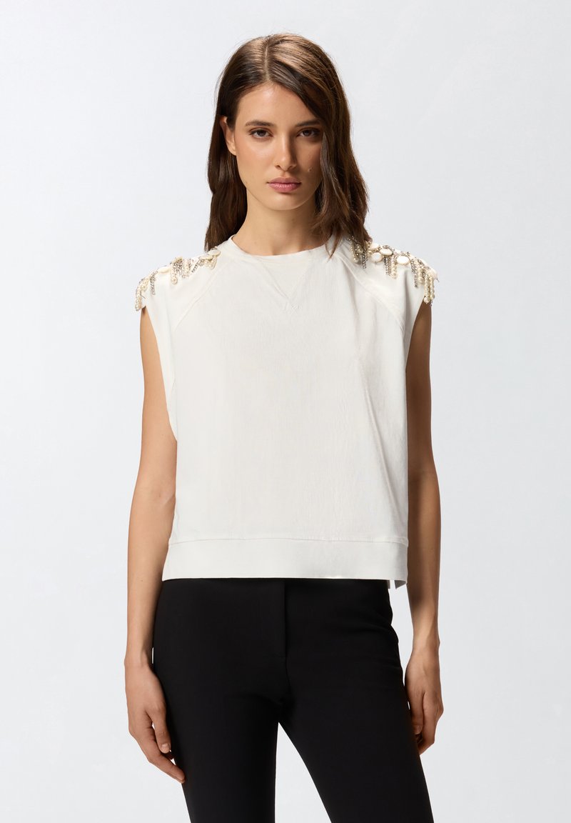 Top blanco sin mangas con tela texturizada, dobladillo acanalado y adornos decorativos en los hombros. Combinado con pantalones ajustados negros.