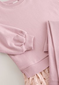 Weicher rosa Strickpullover mit gerippten Bündchen und Saum, kombiniert mit gerippten rosa Leggings und einem floralen Rock darunter.