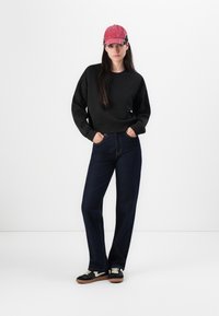 Felpa nera, jeans blu scuro, cappellino rosso e scarpe nere con lacci bianchi. La felpa ha un collo rotondo e una vestibilità ampia.