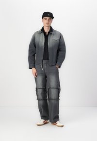 Denim jacket and pants set in gray with a faded effect, frayed edges on pants, and a logo on the jacket. Accented with a black cap and sneakers.