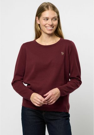 Bordeaux pulover z dolgimi rokavi, okroglim izrezom, rebrastimi manšetami in gladko teksturo. Ima majhen vezen akcent na prsih.