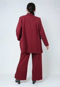 Burgundy tailored suit with a long blazer and wide-leg trousers. Smooth fabric with subtle texture, featuring side slits at the pants.