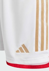 White sports shorts with gold three-stripe design, red trim, and embroidered gold logo on the left side. Smooth, lightweight fabric.