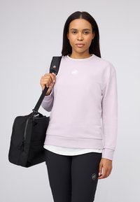 Kvinna med långt mörkt hår bär en ljuslila sweatshirt och svarta byxor, bärande en svart axelväska mot en ljus bakgrund.