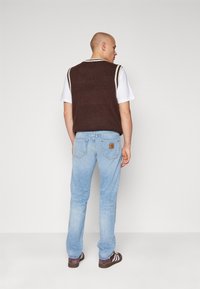 Vesta tricotată maro peste un tricou alb, blugi din denim albastri și pantofi maro cu dungi albe; detaliu de hardware vizibil pe blugi.