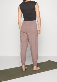 Heller rosa gerippte Jogginghosen mit einem gerafften Bund und umschlägigen Knöcheln, kombiniert mit einem schwarzen ärmellosen Oberteil. Barfuß auf einer Yogamatte.