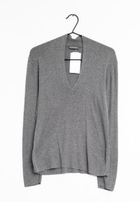 Pull long gris à manches longues avec un décolleté en V profond et des poignets côtelés. Le tissu semble doux avec une texture lisse, suspendu à un crochet noir.