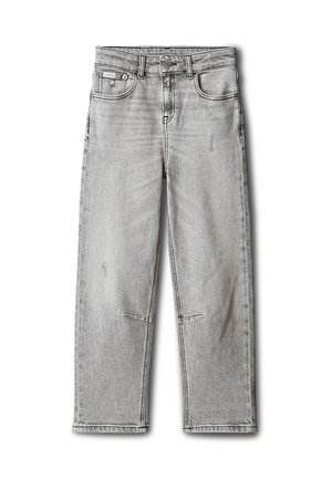 Enfant aux cheveux bruns mi-longs, portant une veste matelassée noire, un t-shirt blanc, un jean gris et des baskets blanches sur un fond gris.