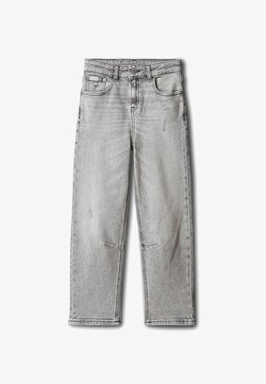 Kind met schouderlang bruin haar, gekleed in een zwarte doorgestikte jas, wit T-shirt, grijze spijkerbroek en witte sneakers op een grijze achtergrond.