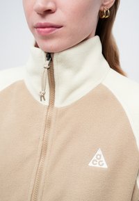 Fleeces jaka ar krēmkrāsu un bēša krāsu bloku dizainu, pus-izvilkšanas aizdari un trīsstūrainu izšūtu logotipu uz krūšu.