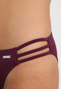 Burgundsko dno bikinija z gladko teksturo, izrezanima obliko in dvojno stransko trakovi, ter srebrnim logotipom.