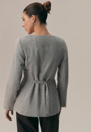 Femeie cu părul prins într-un coc, purtând un sacou gri cu model herringbone și o curea în spate, și pantaloni negri, stând sprijinită de un fundal bej simplu.