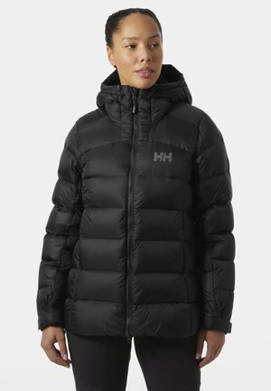 Donna con giacca imbottita nera trapuntata con cappuccio, chiusa fino al collo, in piedi davanti a uno sfondo chiaro e uniforme.