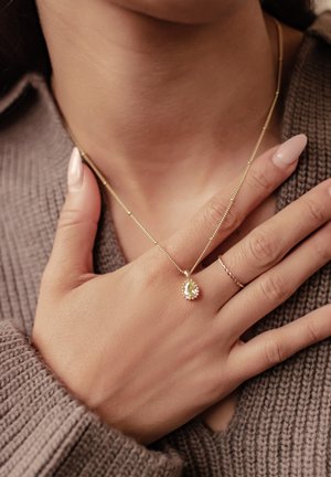 Gold necklace with a pear-shaped gemstone pendant, delicate chain, worn with a thin gold ring on a hand against a textured sweater.