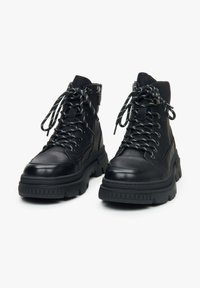 Zwarte leren hoge schoenen met dikke zolen en ronde neuzen. Voorzien van zwart-witte gestreepte veters, een zijrits en textuurdetails.