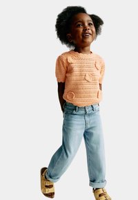 Crochet top v broskvové barvě s květinovými detaily, v kombinaci se světlými modrými džíny a hnědými sandály. Měkká textura, žebrovaný výstřih, krátké rukávy.