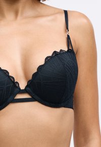 Femeie îmbrăcată cu un sutien din dantelă neagră cu modele geometrice, bretele ajustabile și benzi orizontale duble în față, dedesubtul cupelor.