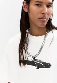 Hvid t-shirt med et sort bilgrafik, båret med en tyk sølvkædehalskæde. Modellen har langt hår og et neutralt udtryk.