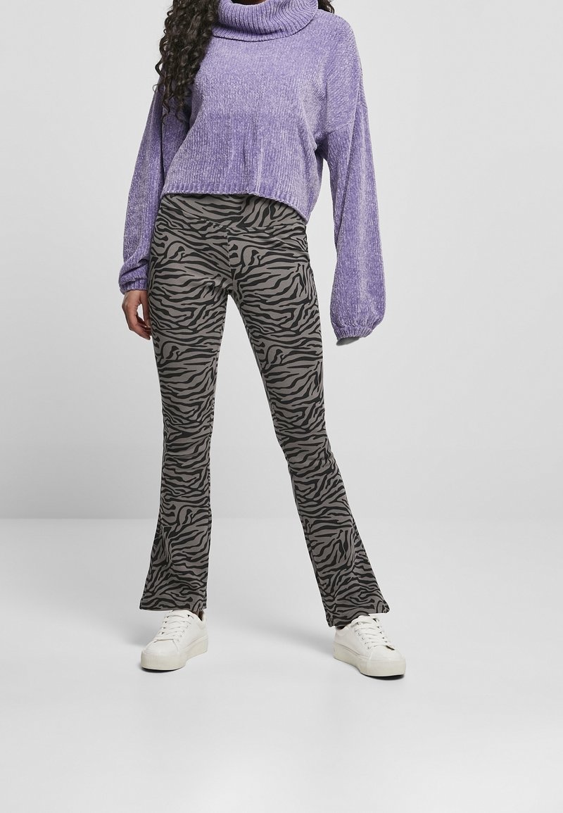 Femme vêtue d'un pull à col roulé violet, pantalon évasé à motifs zèbre gris et baskets blanches, debout devant un fond clair uni.