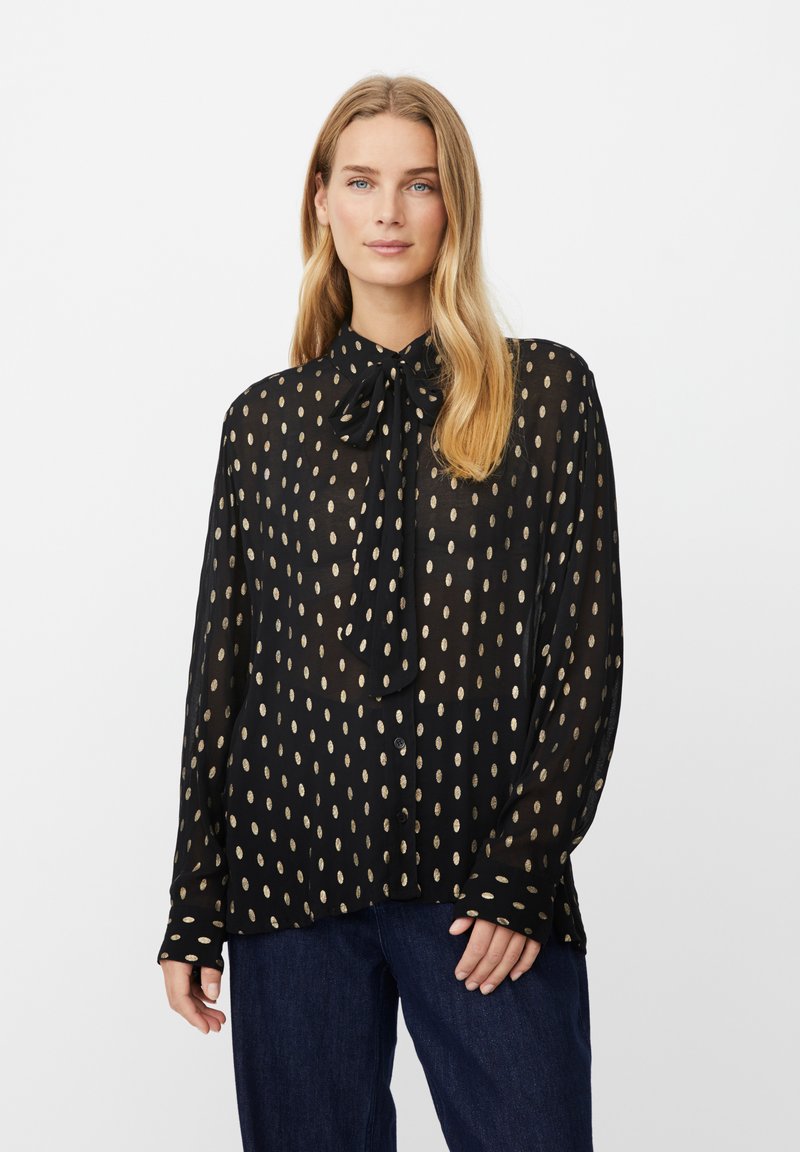 Schwarze, transparente Bluse mit einer großen Schleife am Hals, die goldfarbene ovale Punkte aufweist, lange Ärmel hat und eine geknöpfte Vorderseite besitzt. Dazu werden Jeans aus Denim getragen.