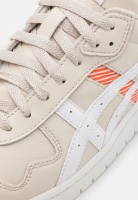 Beige lädersneakers med vita accenter, med texturerade ränder i orange och vitt. Inkluderar beiga snören och framträdande sömnadsdetaljer.