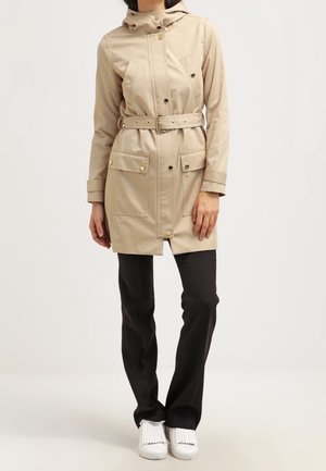 Frau trägt einen beigen Regenmantel mit Gürtel und Druckknöpfen an den Taschen, schwarze Hose und weiße Slipper, steht vor einem schlichten hellen Hintergrund.