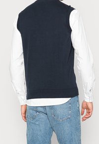 Chaleco de punto azul marino con escote redondo, bajo acanalado y diseño sin mangas, usado sobre una camisa blanca de manga larga y jeans azules.