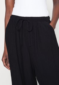 Pantalons larges plissés noirs en tissu texturé. Dotés d'une taille élastique avec cordon de serrage et de poches latérales.