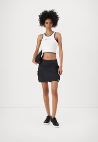 Fehér cropped tank top fekete díszítéssel és felirattal, fekete sportszoknyával párosítva, amely húzózsinórral és zsebekkel rendelkezik. Fekete sportcipő.