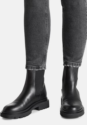 Stivaletti alla caviglia in pelle nera con finitura liscia, inserti elastici ai lati e suole spesse in gomma, abbinati a jeans grigi slavati con effetto usurato.