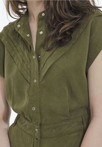 Mujer con una blusa sin mangas de botones de color verde oliva, con detalles plisados y un cinturón a juego en la cintura.