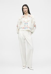 Γυναίκα που στέκεται φορώντας λευκό παντελόνι με φαρδιά πόδια, λευκό πουκάμισο με πολύχρωμη κεντητή λουλουδένια διακόσμηση και δαντελωτές λεπτομέρειες, και λευκά μυτερά τακούνια.