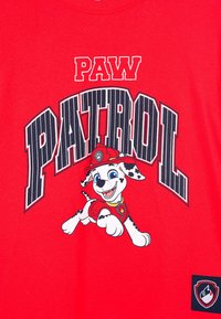 Czerwona koszulka z rysunkowym dalmatyńczykiem w hełmie strażackim i kamizelce, z dużym napisem "PAW PATROL" powyżej.