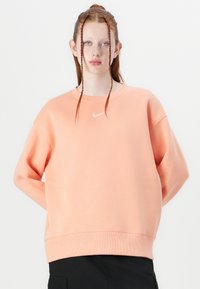 Sweatshirt oversized em pêssego com um decote redondo, bainha canelada e logotipo branco da Nike no peito. O material parece suave e liso.