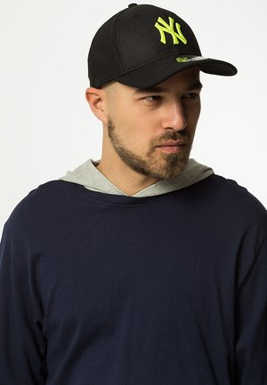Schwarze Baseballmütze mit einem Netzdesign, versehen mit einem leuchtend gelben NY-Logo. Getragen mit einem navyblauen Langarmshirt und einer grauen Kapuzenjacke.
