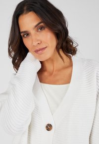 Cardigan de punto blanco con escote en V y patrón texturizado. Cuenta con un solo botón marrón en la parte delantera, llevado sobre una blusa clara.