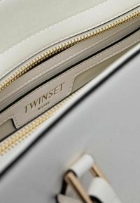 Interno di una borsa in pelle bianca con cerniera dorata e fodera nera. Etichetta visibile che recita "TWINSET MILANO." Texture liscia con design strutturato.
