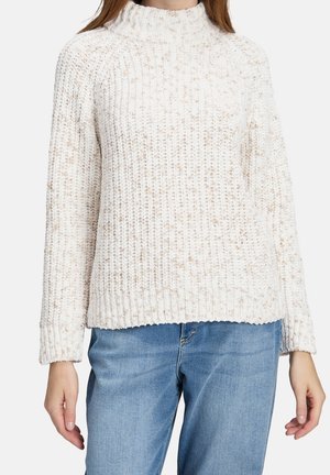 Femme portant un pull en maille épaisse de couleur crème à manches longues et un jean denim bleu clair, debout devant un fond uni.
