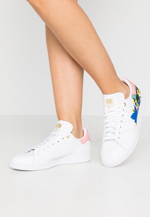 Nogi w białych tenisówkach Adidas Stan Smith z różowymi panelami na pięcie i kolorowymi kwiatowymi wzorami po bokach, stojące na jasnej podłodze.