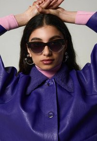 Veste en cuir violet avec un col en fourrure, superposée à un col roulé rose. Le modèle porte des lunettes de soleil dégradées noires et des boucles d'oreilles rondes en argent.