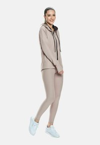 Woman smiling, wearing a beige hooded zip-up jacket, matching leggings, and white sneakers, standing sideways on a white background.