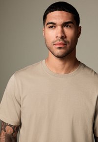 Junger Mann mit kurzen dunklen Haaren und tätowiertem Arm, der ein beiges T-Shirt trägt und leicht nach rechts blickt, vor einem schlichten grauen Hintergrund.