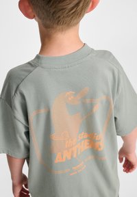 Ljusgrön bomullströja med en orange grafik på baksidan med texten "the stadium ANTHEMS" och en tecknad karaktärsdesign.