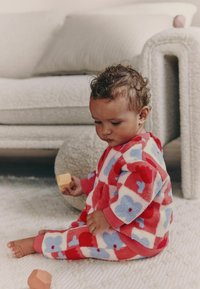 Fleece-asuste, jossa on punainen ja vaaleansininen kukkakuosi, ribbineulokset hihansuissa ja kirkas värimaailma; lapsi pitää lelupalikkaa.