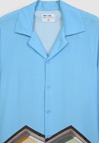 Camicia azzurra a maniche corte con colletto a punta, caratterizzata da una stampa geometrica in colori più scuri all'orlo e due bottoni lungo la parte anteriore.