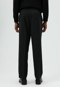 Zwarte op maat gemaakte broek met een rechte pasvorm, voorzien van twee achterzakken en een gestructureerde afwerking, gedragen met zwarte schoenen.