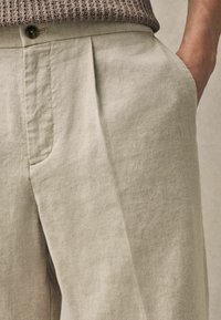 Pantalons en lin beige clair avec une coupe décontractée, des plis verticaux et des poches latérales. Comprend une fermeture à un seul bouton au niveau de la taille.