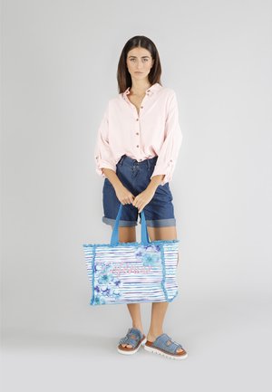 Sac fourre-tout floral bleu avec un motif rayé, des bords effilochés et des lettres roses. Modèle portant une chemise rose clair et un short en denim.