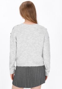 Kobieta z długimi brązowymi włosami, ubrana w jasnoszary puszysty sweter i ciemnoszary plisowany spódnicę, stoi tyłem na białym tle.