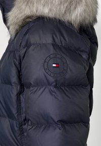 Nærbillede af en marineblå puffervestærme med pelskant og et rundt Tommy Hilfiger-logo-patch.